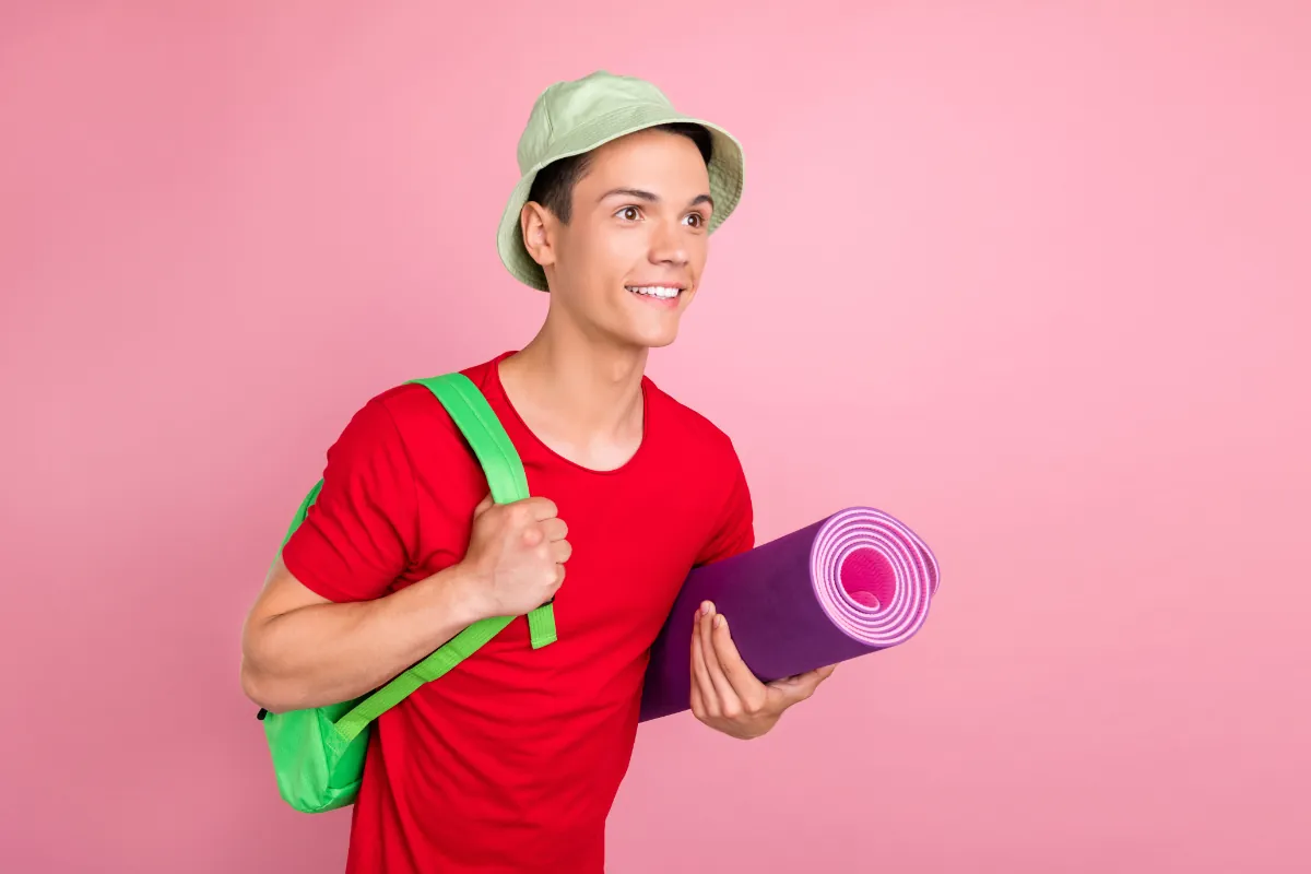 Un jeune homme en tee-shirt rouge, sac au dos et tapis de yoga roulé sous le bras, sur fond rose