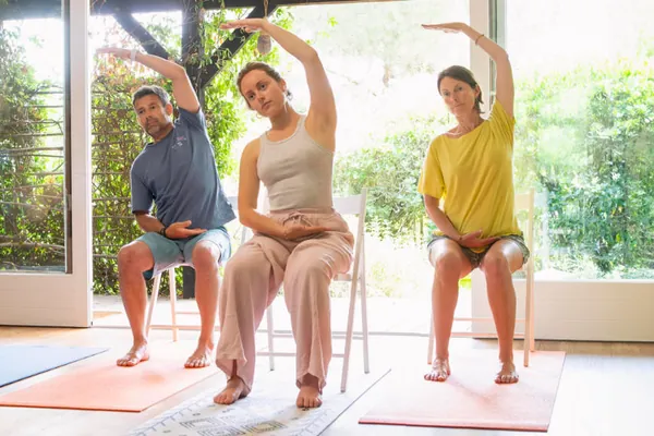 Trois pratiquants en yoga sur chaise, en flexion latérale devant la baie vitrée